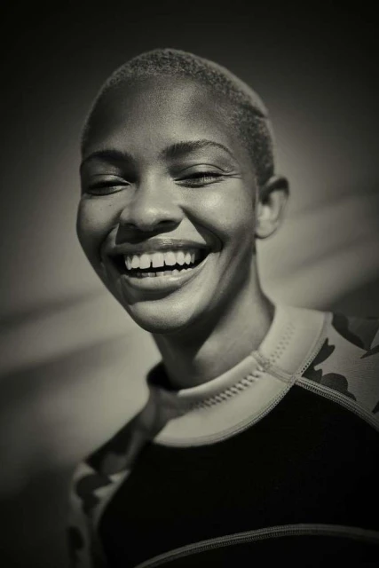 Monochromatic close-up of a smiling adult female with a buzzcut, wearing a patterned rash guard, in front of a neutral background.