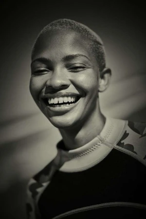 Monochromatic close-up of a smiling adult female with a buzzcut, wearing a patterned rash guard, in front of a neutral background.