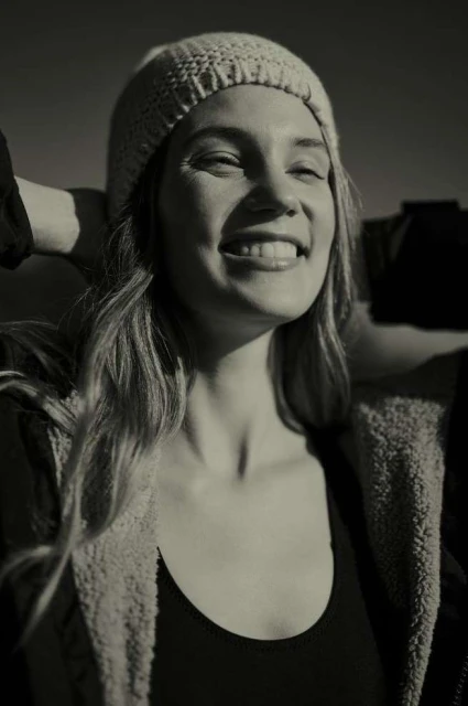 Close-up shot of an adult woman smiling with a beanie on her head; shot in black and white outdoors with her hands behind her head.