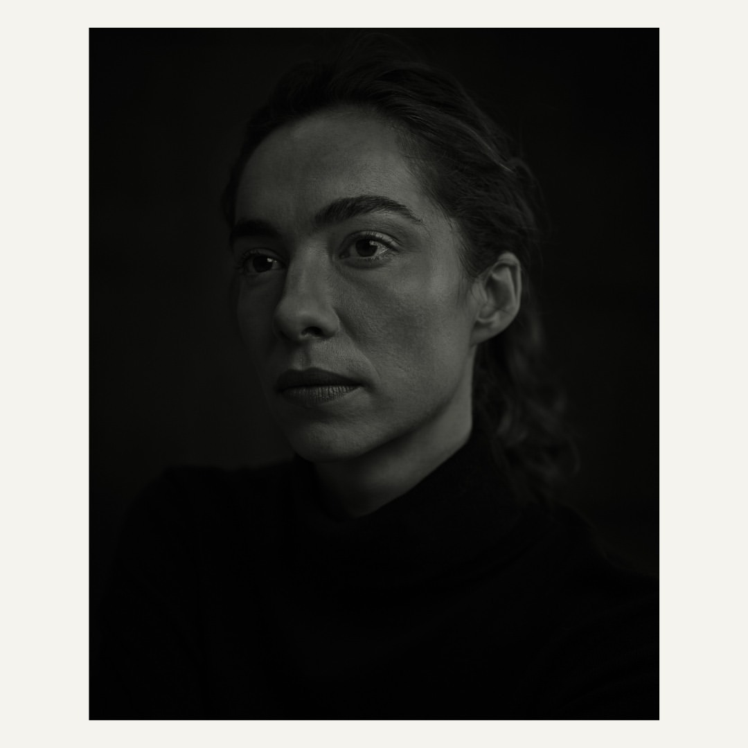 Monochrome portrait of a woman with dark hair in a black turtleneck against a dark background, emphasizing light and shadow.