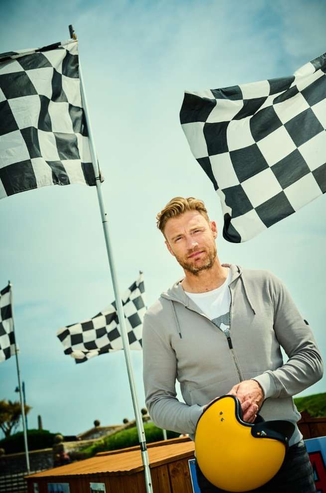 Adult male poses outdoors with a yellow helmet in hand, set against checkered racing flags and a partly cloudy blue sky.