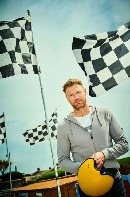 Adult male poses outdoors with a yellow helmet in hand, set against checkered racing flags and a partly cloudy blue sky.