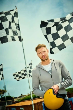Adult male poses outdoors with a yellow helmet in hand, set against checkered racing flags and a partly cloudy blue sky.