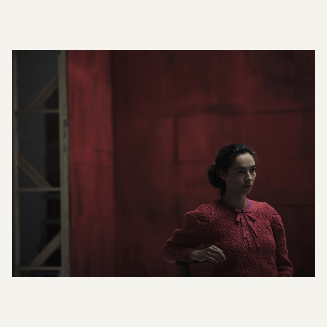 Woman in a red sweater sits in a room with red walls and window frame. Portrait photograph in indoor setting with soft lighting.