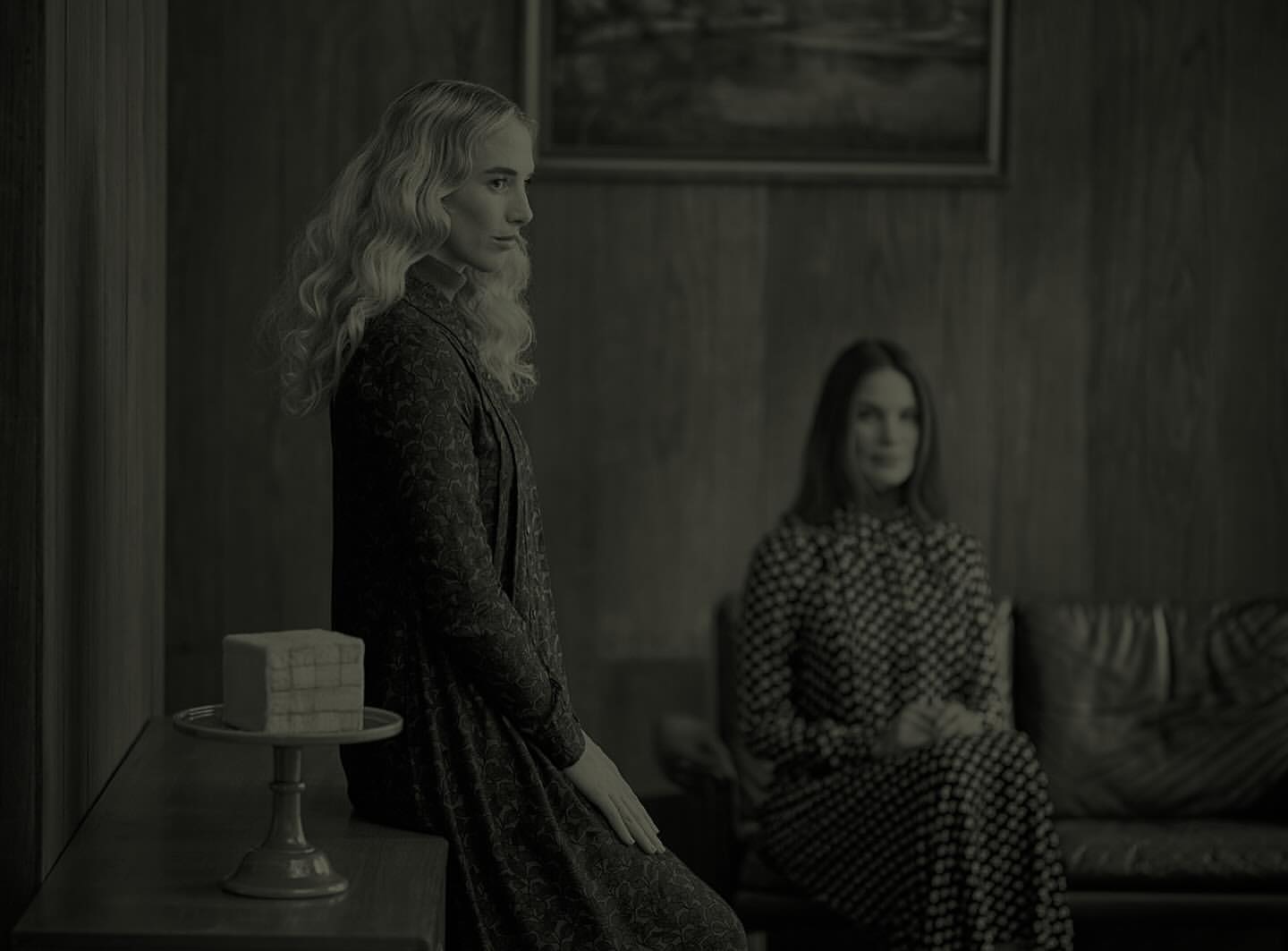 Monochrome portrait of two adult women in vintage dresses, one seated, the other standing near a cake, in an indoor setting.