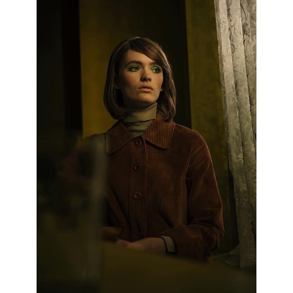 Young woman with green eye shadow and a brown corduroy jacket poses in a dimly lit indoor setting, with patterned curtains in the background.