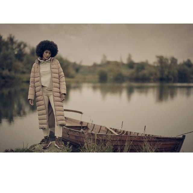 Adult female in a long beige coat standing on the shore of a lake with a small wooden rowboat and an overcast sky in the background.