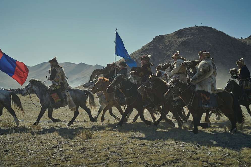 Horsemen in fur coats ride across a plain towards mountains under a clear sky, carrying flags and creating a dramatic, historic scene.