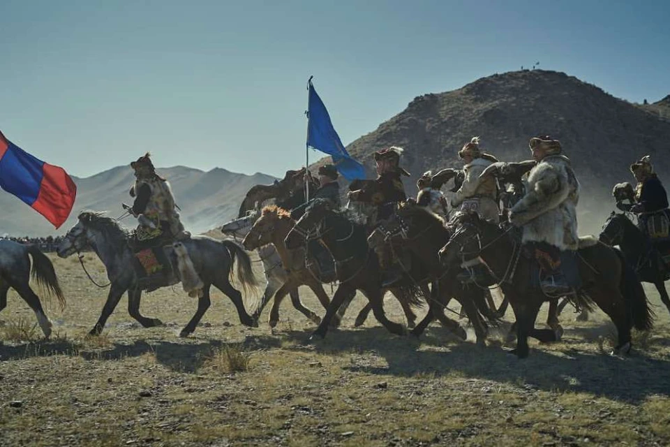 Horsemen in fur coats ride across a plain towards mountains under a clear sky, carrying flags and creating a dramatic, historic scene.