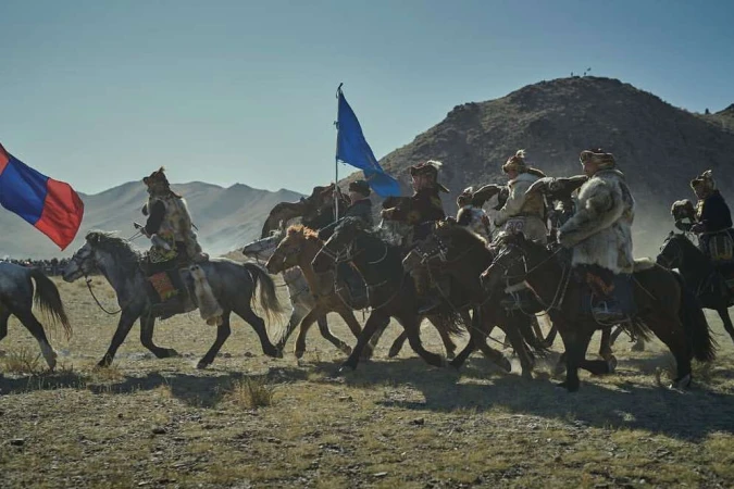 Horsemen in fur coats ride across a plain towards mountains under a clear sky, carrying flags and creating a dramatic, historic scene.