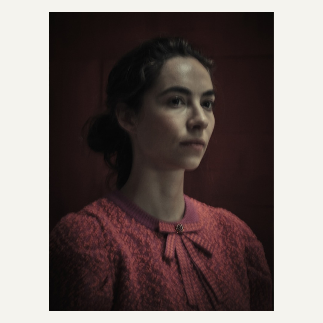 Portrait of a young woman with her hair in a bun, wearing a pink tweed cardigan with a bow, against a red-toned background.