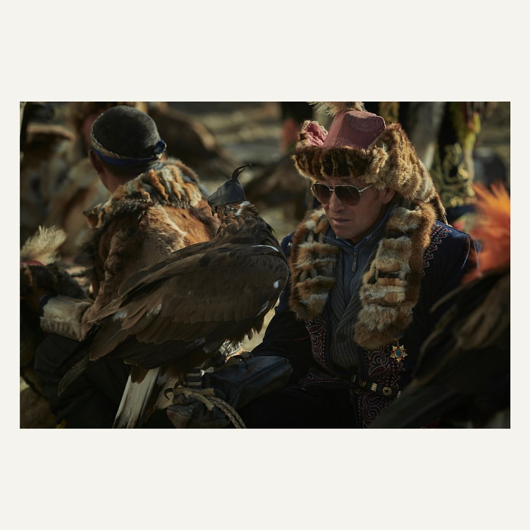 Mongolian eagle hunter in traditional clothing with his trained golden eagle perched on arm, sunglasses, fur hat, in outdoor setting.