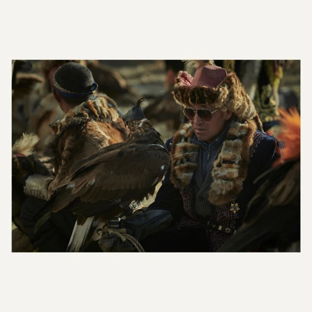 Mongolian eagle hunter in traditional clothing with his trained golden eagle perched on arm, sunglasses, fur hat, in outdoor setting.