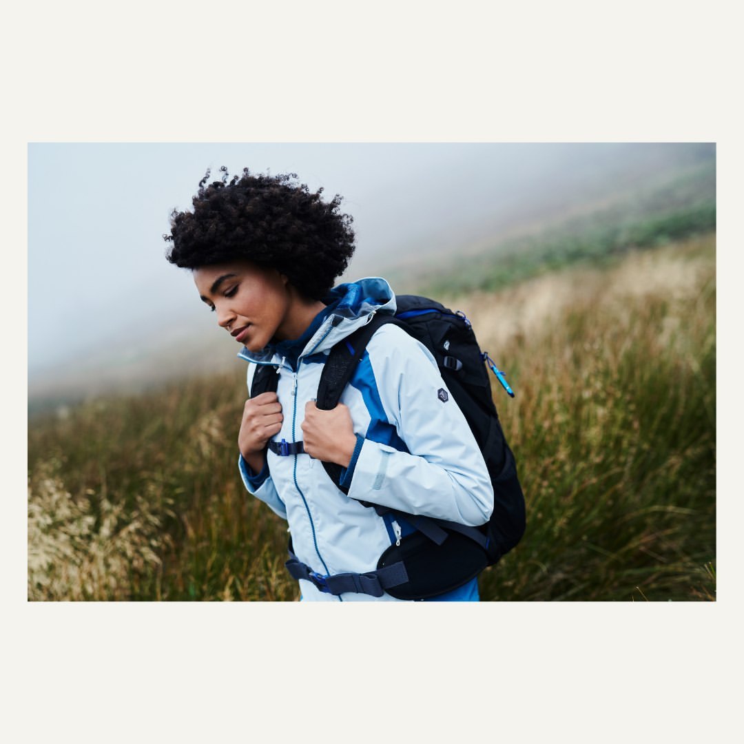 Woman hiking on a grassy hill in a light blue jacket and black backpack. Fog is rolling in over the green grass.
