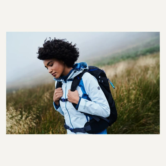 Woman hiking on a grassy hill in a light blue jacket and black backpack. Fog is rolling in over the green grass.