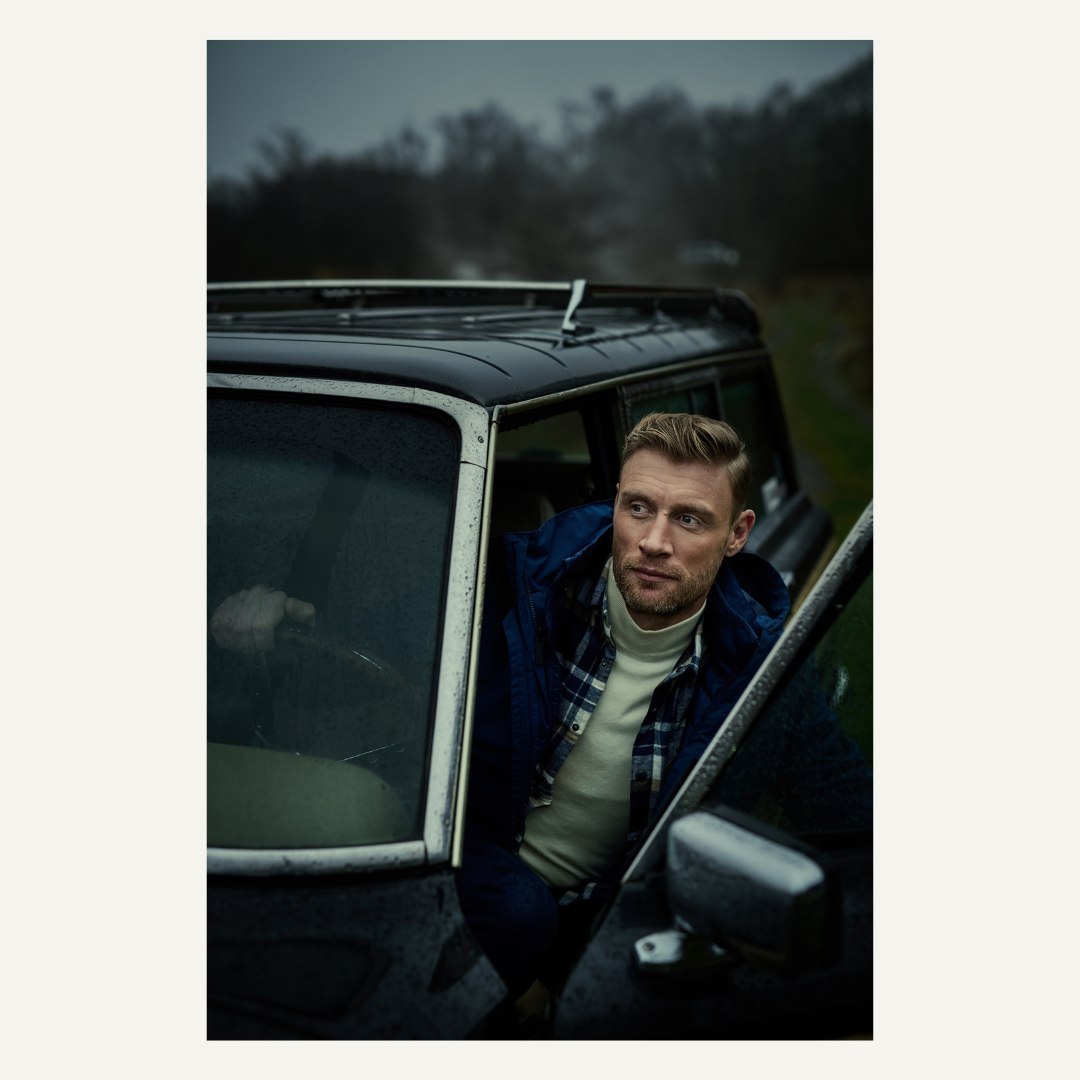 A man stands by an old black SUV in a field, wearing a flannel shirt and turtleneck sweater under a navy blue coat, gazing off into the distance.