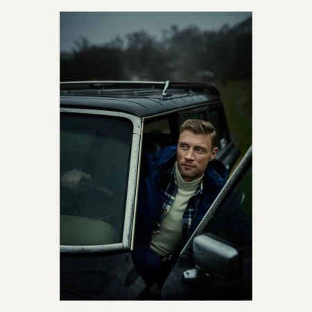 A man stands by an old black SUV in a field, wearing a flannel shirt and turtleneck sweater under a navy blue coat, gazing off into the distance.