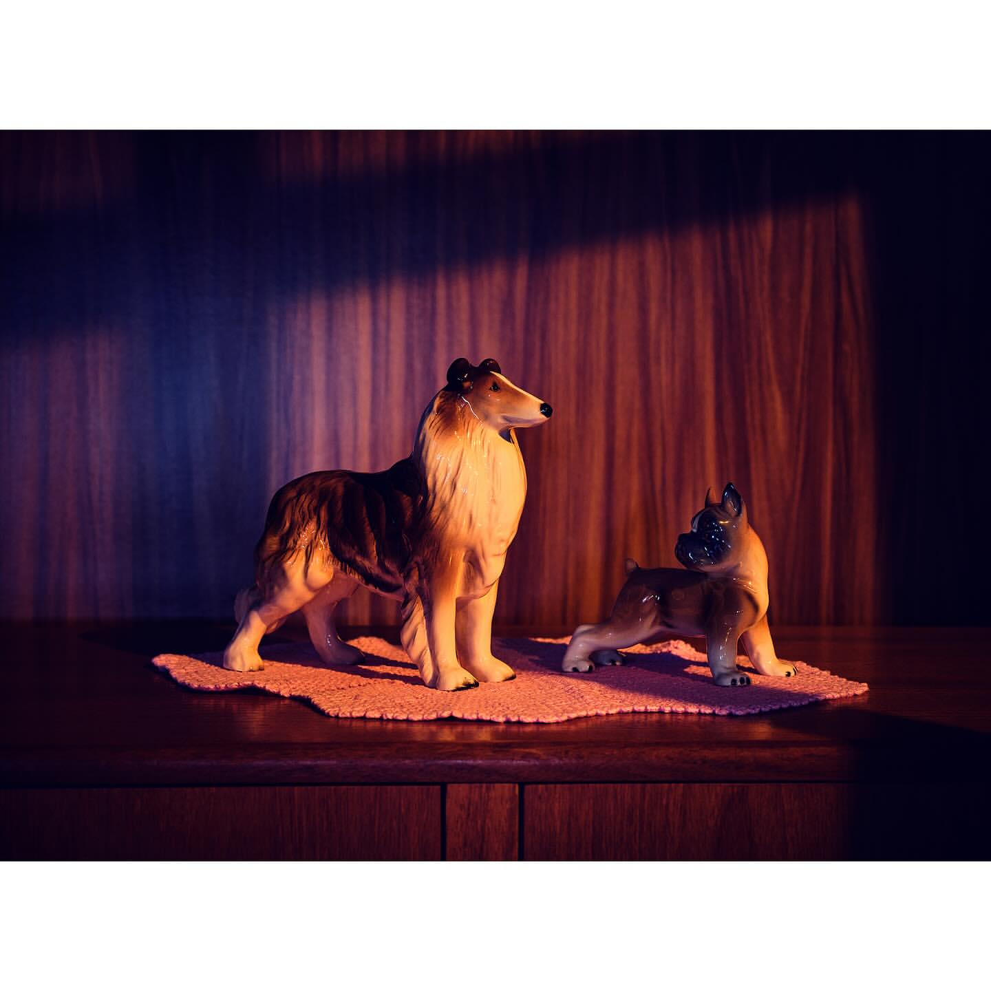 Vintage figurine display featuring a collie and a boxer dog on a pink crocheted doily atop a dark wooden cabinet with patterned backdrop.