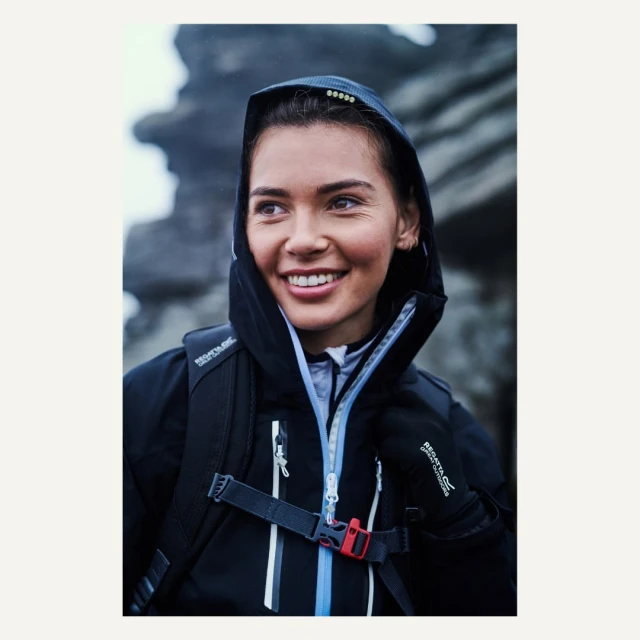 Smiling woman in a dark hooded waterproof jacket and backpack in an outdoor environment with grey rock formations in the background.