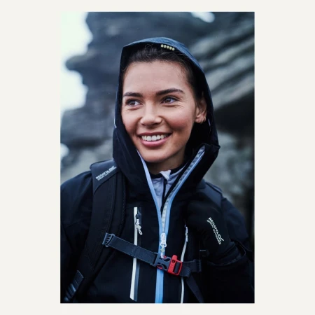 Smiling woman in a dark hooded waterproof jacket and backpack in an outdoor environment with grey rock formations in the background.