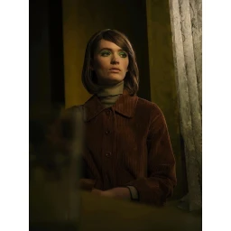 A woman with short brown hair and green eyeshadow wearing a brown corduroy jacket and turtleneck sitting indoors.