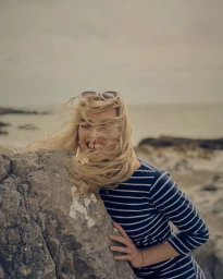 Smiling blonde woman with hair blowing across her face leaning against a large rock on a beach.
