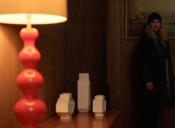 Woman in dark jacket and beanie standing in a wood paneled room with a red lamp and white vases by David Short.