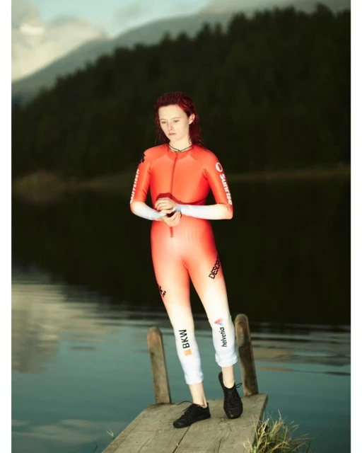 Female fashion model poses in a sporty red gradient jumpsuit on a wooden pier with lake and mountains background in an outdoor setting.