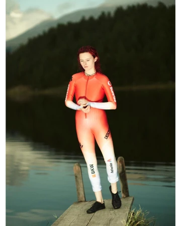 Female fashion model poses in a sporty red gradient jumpsuit on a wooden pier with lake and mountains background in an outdoor setting.