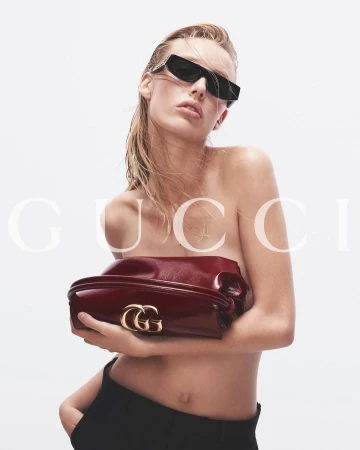 Model wearing dark sunglasses holding a glossy dark red patent leather clutch bag against a plain white background.