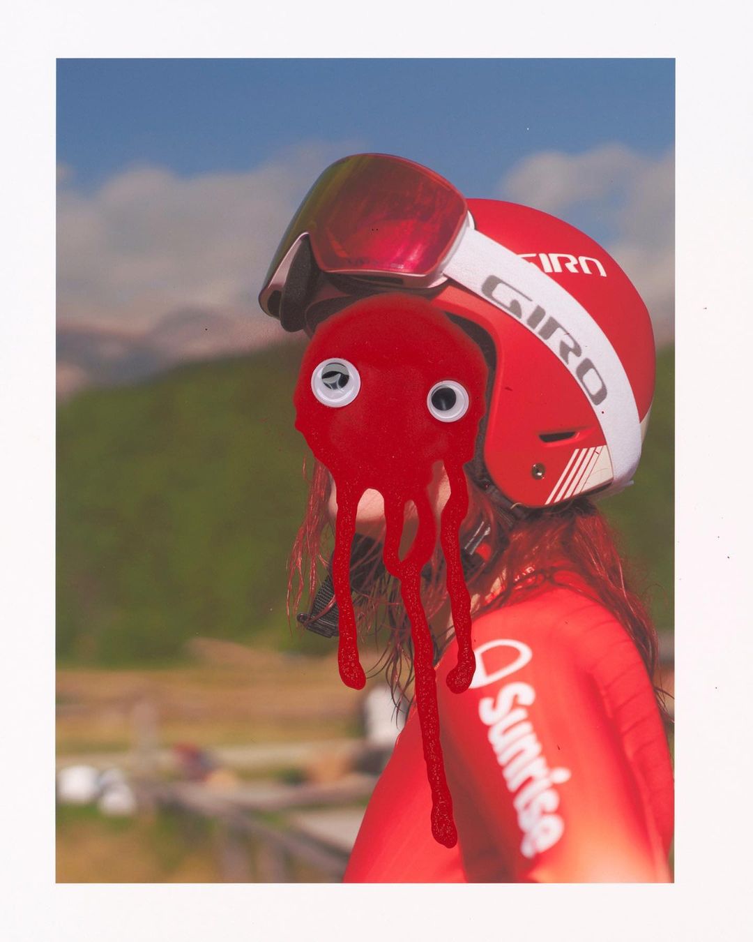 A surreal image of a person wearing a red Giro ski helmet with a red octopus attached, set against a mountain landscape under a bright sky.