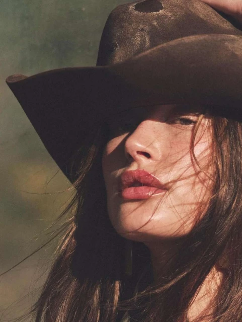 Close-up portrait of a woman wearing a brown cowboy hat with windblown brown hair, standing outdoors against a soft, blurred background.