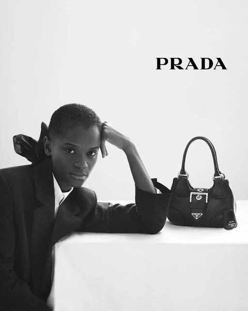 Black-and-white fashion shot features an adult woman wearing a black blazer posing on a white surface next to a black Prada handbag.