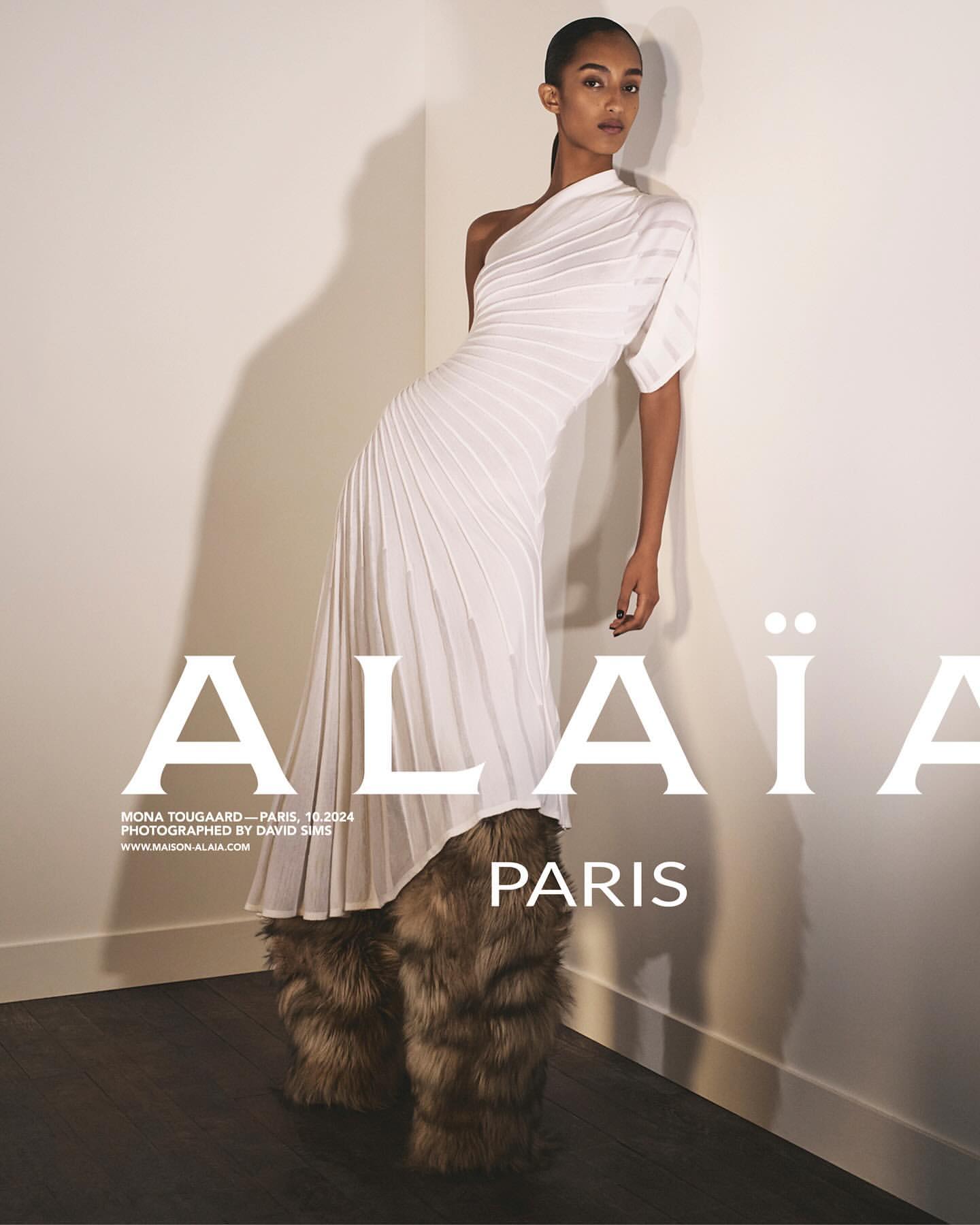Editorial fashion shot of a woman in a white one-shoulder gown and fur-like boots posing in front of a white wall with brand typography.