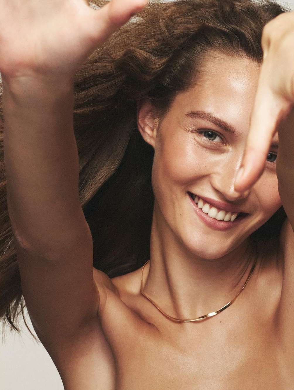 Close-up studio shot of smiling woman with long brown hair framing her face, arms raised with hands making a square, and wearing a gold necklace.