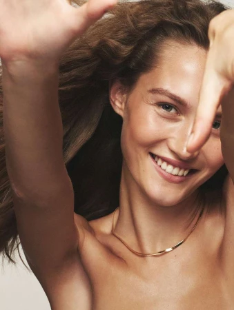 Close-up studio shot of smiling woman with long brown hair framing her face, arms raised with hands making a square, and wearing a gold necklace.