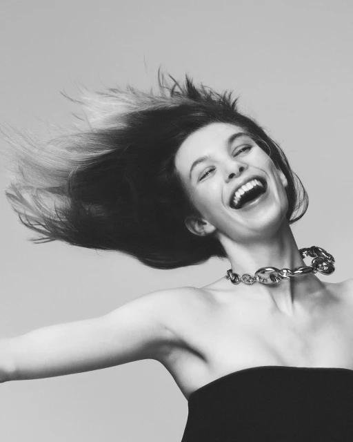 Portrait of a young woman with open mouth, wearing a black strapless top and silver chain link necklace against a plain background.