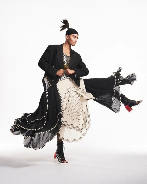 Fashion-forward woman in an asymmetric black jacket and patterned skirt strikes a dynamic pose in a studio setting with a solid white backdrop.