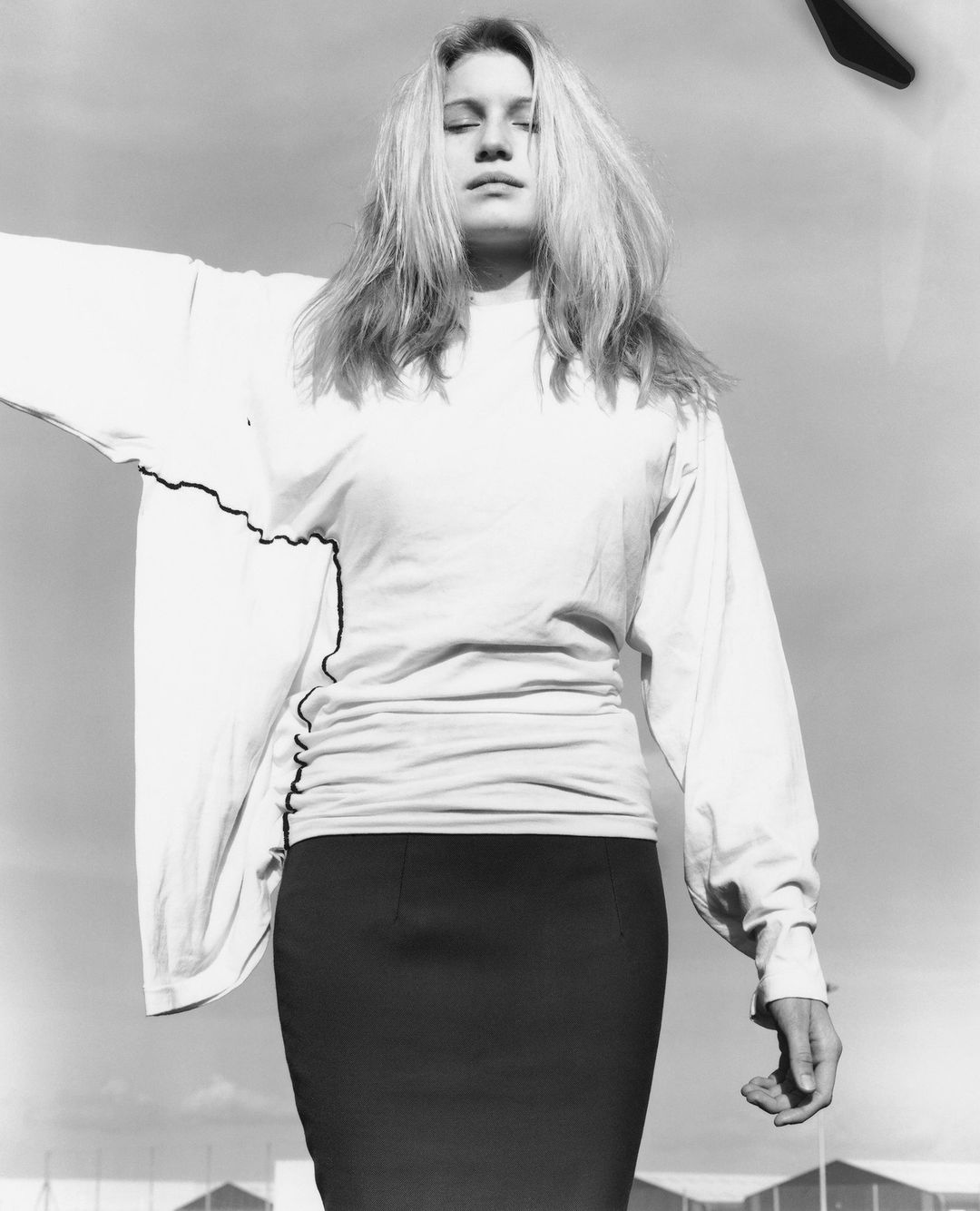 Monochrome portrait of a blonde woman in a white long-sleeve top and dark skirt against an overcast sky, photographed by David Sims.