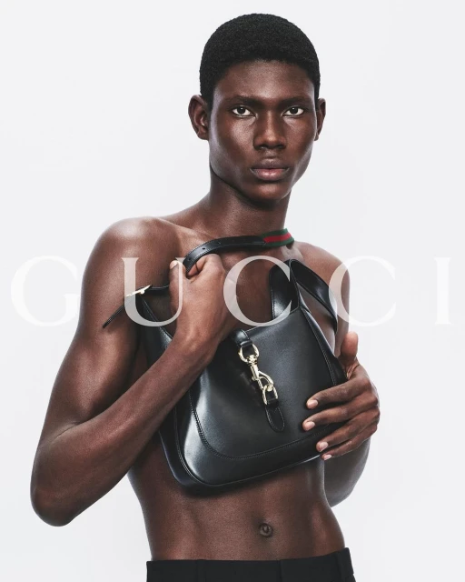 A fashion shot features a Black male model, bare-chested, holding a black leather Gucci Jackie 1961 bag against a bright white backdrop.