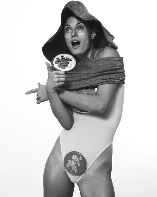 Monochrome image of a woman in a swimsuit and hat, holding decorative plates, pointing. Shot in a photo studio with a white backdrop.