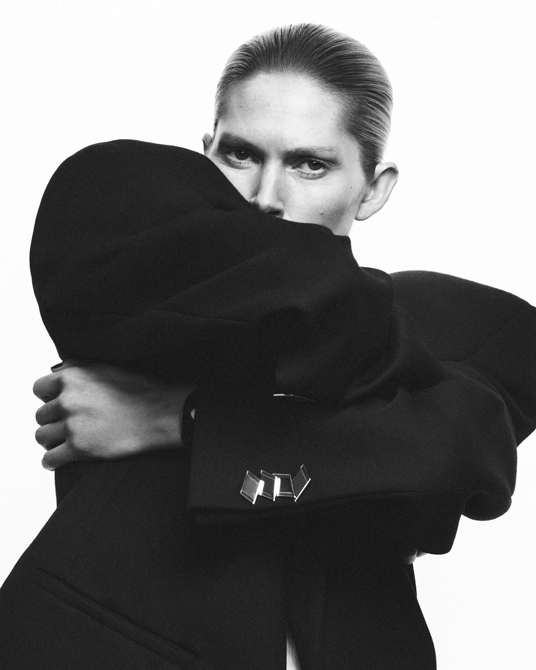 Black and white fashion portrait of an androgynous adult model wearing a sculptural black jacket with metallic cufflink details indoors.