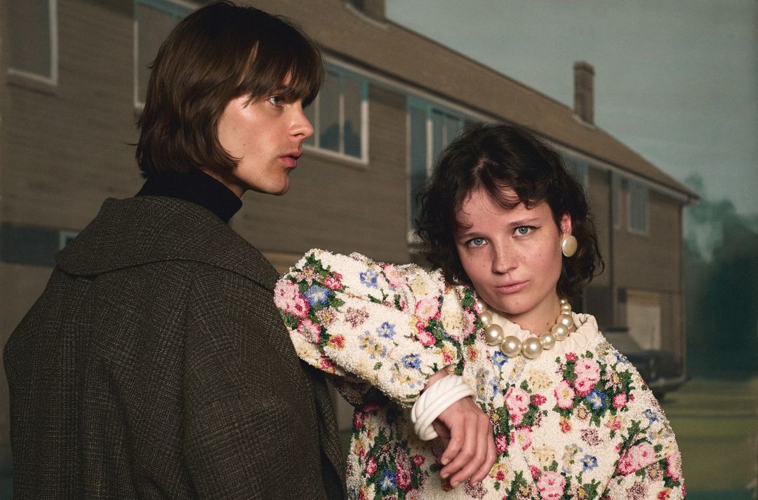 A couple poses in front of a house, woman in a floral sweater and pearls, man in a coat, creating an editorial fashion image.