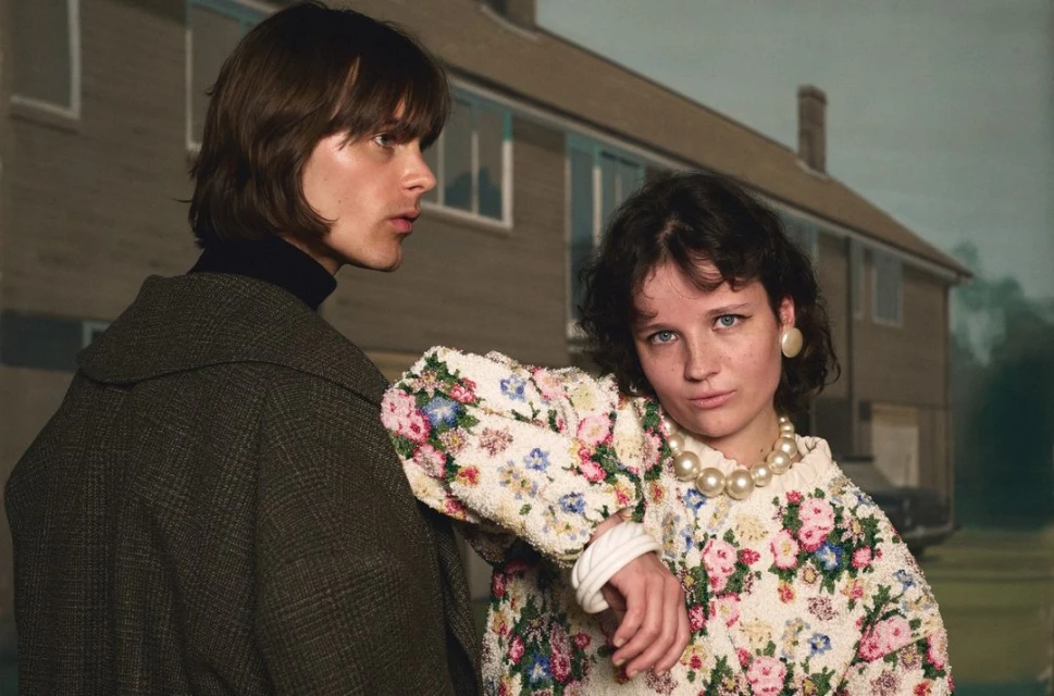 A couple poses in front of a house, woman in a floral sweater and pearls, man in a coat, creating an editorial fashion image.