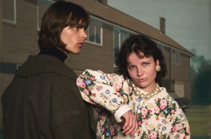 A couple poses in front of a house, woman in a floral sweater and pearls, man in a coat, creating an editorial fashion image.