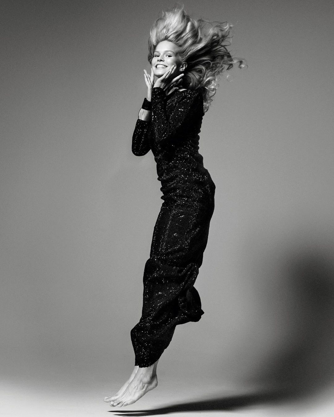 Monochrome full-length portrait of a smiling blonde woman jumping in a long sequined gown against a grey studio backdrop.