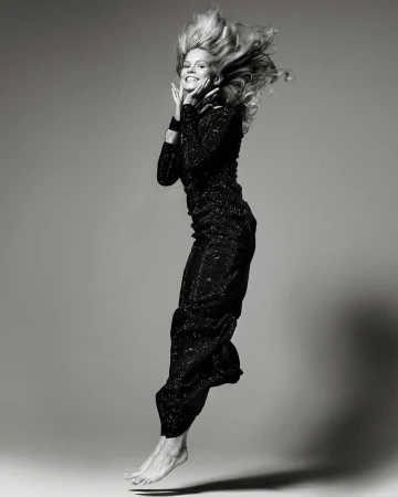 Monochrome full-length portrait of a smiling blonde woman jumping in a long sequined gown against a grey studio backdrop.