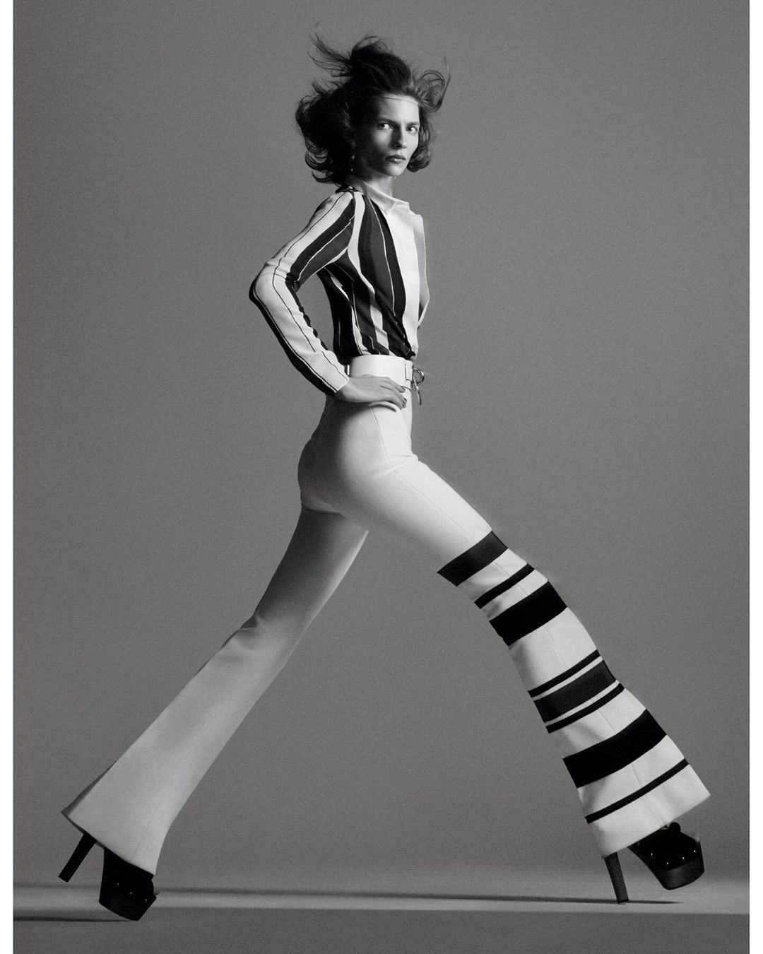 A fashion shot in black and white presents a woman with windblown hair wearing a striped top and striped bell-bottom pants, posing in a studio.