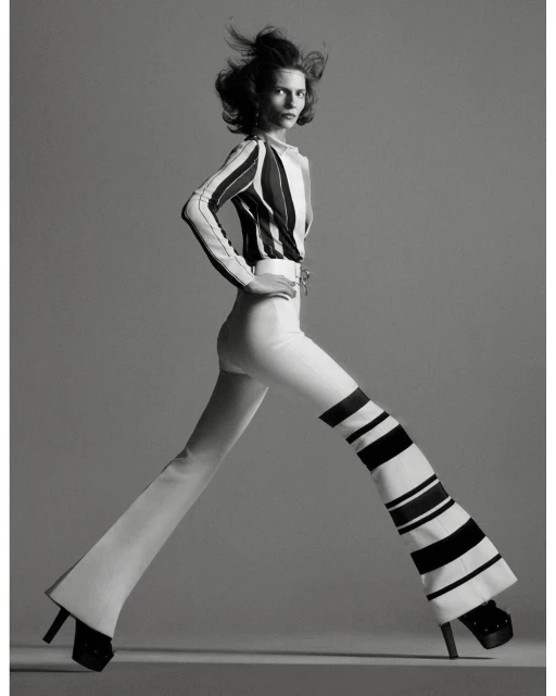 A fashion shot in black and white presents a woman with windblown hair wearing a striped top and striped bell-bottom pants, posing in a studio.