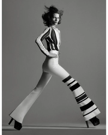 A fashion shot in black and white presents a woman with windblown hair wearing a striped top and striped bell-bottom pants, posing in a studio.